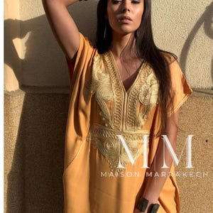 May include: A woman wearing a long, mustard yellow kaftan with gold embroidery. The kaftan has a V-neckline and short sleeves. The woman is wearing a black and white bracelet on her left wrist. The text "MMM MAISON MARRAKECH" is visible at the bottom of the image.