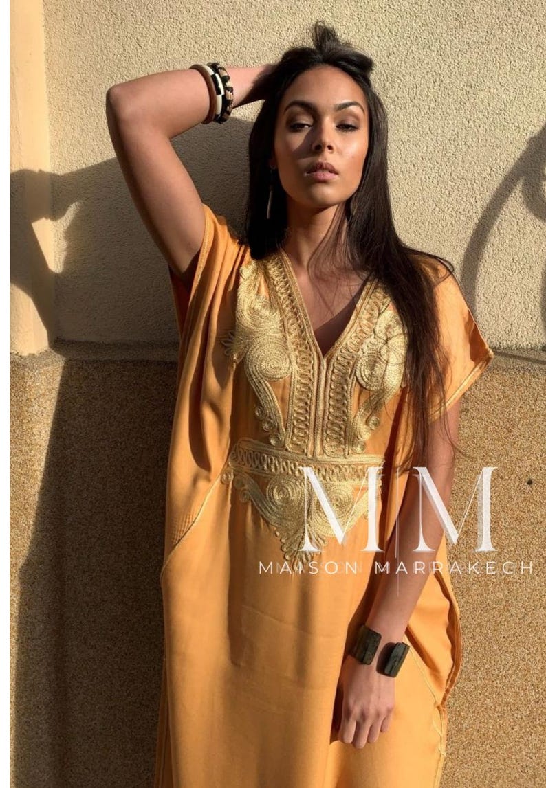 May include: A woman wearing a long, mustard yellow kaftan with gold embroidery. The kaftan has a V-neckline and short sleeves. The woman is wearing a black and white bracelet on her left wrist. The text "MMM MAISON MARRAKECH" is visible at the bottom of the image.