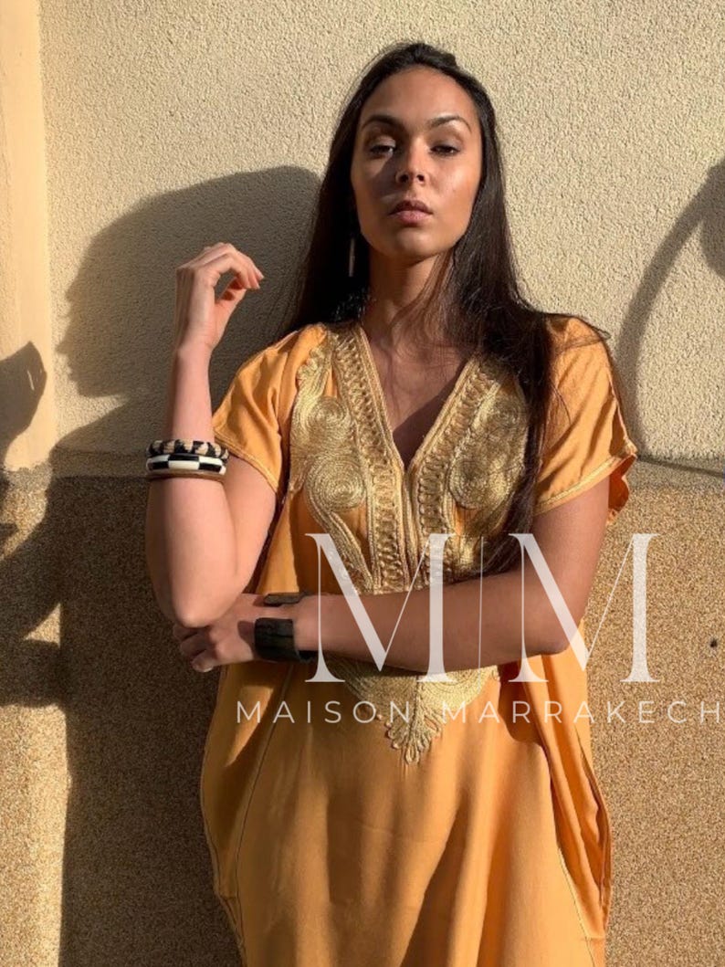 May include: A woman wearing a long, loose-fitting, mustard yellow kaftan with gold embroidery. The kaftan has a V-neckline and short sleeves. The woman is standing in front of a wall with the text "MMM Maison Marrakech" printed on it.