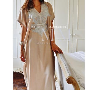 Puede incluir: Un caft&aacute;n largo beige, con cuello en V y mangas cortas. El vestido presenta un intrincado bordado blanco alrededor del escote y el corpi&ntilde;o. El texto "MAISON DE MARRAKECH" es visible en el vestido.