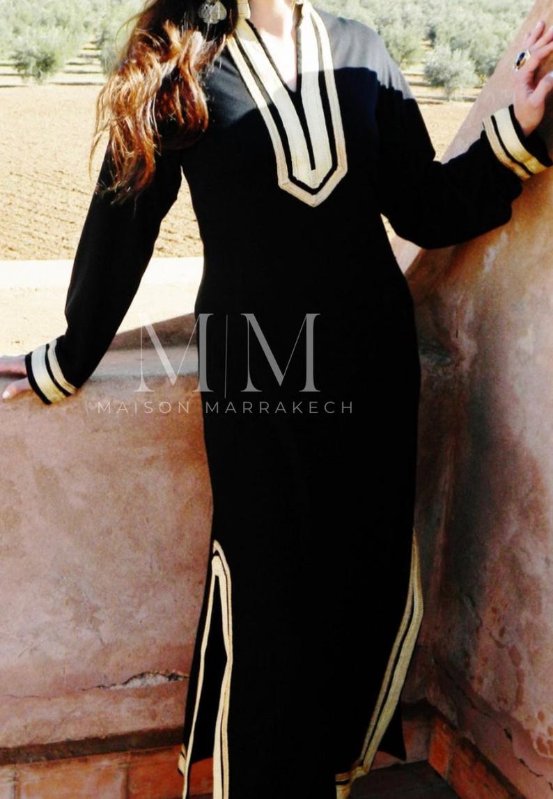 Puede incluir: Un vestido negro de manga larga con ribetes dorados alrededor del escote, los pu&ntilde;os y los lados. El vestido tiene un escote en V y un corte holgado. El texto "MM MAISON MARRAKECH" est&aacute; impreso en la parte delantera del vestido.