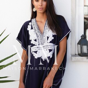May include: A navy blue kaftan with white embroidery. The kaftan has a V-neckline and short sleeves. The embroidery is intricate and features a floral design. The kaftan is made of a soft, flowing fabric. The text "Maison Marrakech" is visible on the bottom of the kaftan.