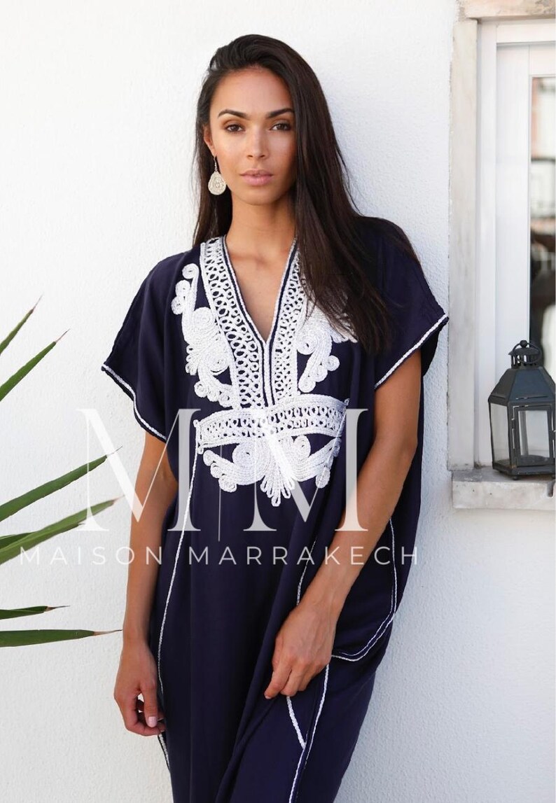 May include: A navy blue kaftan with white embroidery. The kaftan has a V-neckline and short sleeves. The embroidery is intricate and features a floral design. The kaftan is made of a soft, flowing fabric. The text "Maison Marrakech" is visible on the bottom of the kaftan.