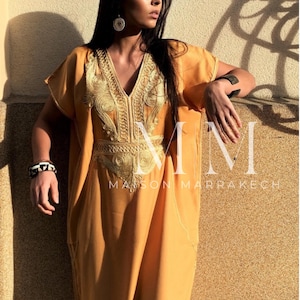 May include: A long, mustard yellow kaftan with gold embroidery. The kaftan has a V-neck and short sleeves. The embroidery features a floral design. The text "MM Maison Marrakech" is visible at the bottom of the image.