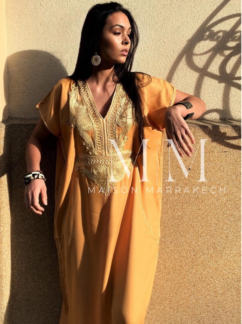May include: A long, mustard yellow kaftan with gold embroidery. The kaftan has a V-neck and short sleeves. The embroidery features a floral design. The text "MM Maison Marrakech" is visible at the bottom of the image.