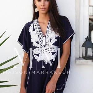 May include: A navy blue kaftan with white embroidery. The kaftan has a V-neckline and short sleeves. The embroidery is intricate and features a floral design. The kaftan is made of a soft, flowing fabric. The text "Maison Marrakech" is visible on the bottom of the kaftan.