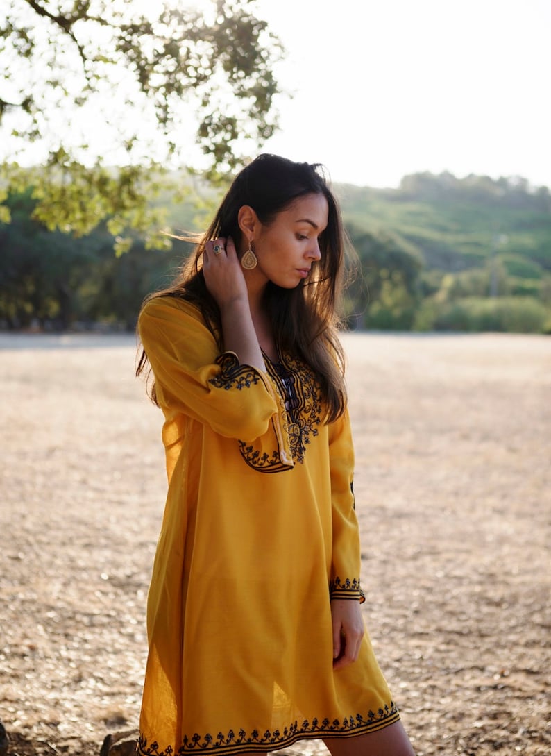 May include: A woman wearing a yellow embroidered dress with black trim. She is standing in a field with a blurred background.