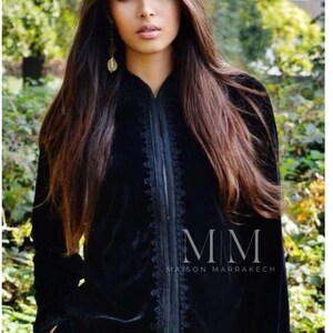May include: A black velvet jacket with intricate embroidery around the collar and front zipper. The jacket has a long, flowing silhouette and features the text "MM Maison Marrakech" on the bottom.