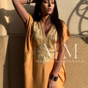 May include: A woman wearing a long, mustard yellow kaftan with gold embroidery. The kaftan has a V-neck and is loose-fitting. The woman is wearing a black and white bracelet on her left wrist and a black bracelet on her right wrist. The text "MM Maison Marrakech" is visible on the bottom of the image.