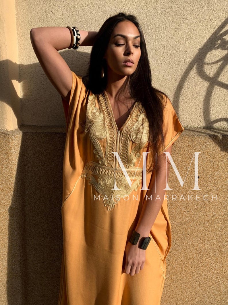 May include: A woman wearing a long, mustard yellow kaftan with gold embroidery. The kaftan has a V-neck and is loose-fitting. The woman is wearing a black and white bracelet on her left wrist and a black bracelet on her right wrist. The text "MM Maison Marrakech" is visible on the bottom of the image.