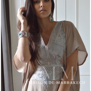 Puede incluir: Un caft&aacute;n beige con intrincados bordados plateados alrededor del escote y el corpi&ntilde;o. El caft&aacute;n tiene mangas cortas y cuello en V. El texto "MAISON DE MARRAKECH" es visible. La modelo lleva una pulsera plateada con piedras de colores.