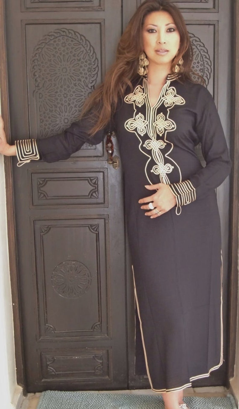 K&ouml;nnte beinhalten: Eine Frau tr&auml;gt ein schwarzes, lang&auml;rmeliges Kleid mit goldener Stickerei. Das Kleid hat einen V-Ausschnitt und einen langen Rock. Die Frau steht vor einer Holzt&uuml;r mit aufwendigen Schnitzereien.