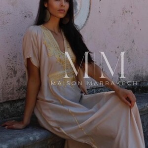 May include: A woman wearing a long, light beige kaftan with gold embroidery and trim. The kaftan has a V-neckline and short sleeves. The woman is sitting on a stone ledge with a pink wall behind her. The text "MM MAISON MARRAKECH" is visible in the image.