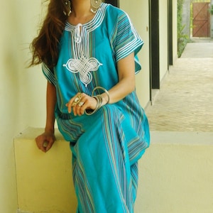 May include: A woman wearing a turquoise and white striped dress with white embroidery. She is sitting on a white ledge with a white wall behind her.