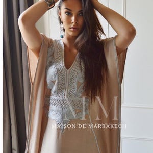 Puede incluir: Un caft&aacute;n beige con intrincados bordados en azul claro alrededor del escote y las mangas. El caft&aacute;n tiene cuello en V y un dise&ntilde;o suelto y fluido. El texto "MAISON DE MARRAKECH" es visible en la prenda.