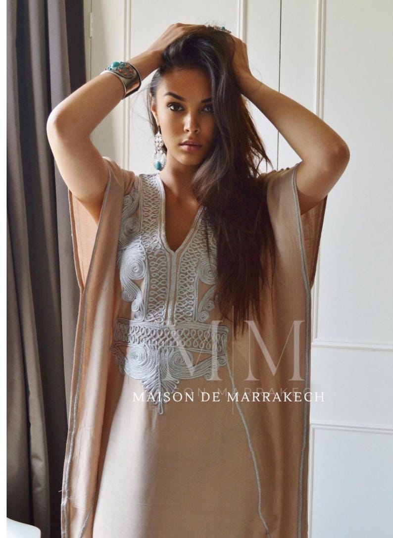 Puede incluir: Un caft&aacute;n beige con intrincados bordados en azul claro alrededor del escote y las mangas. El caft&aacute;n tiene cuello en V y un dise&ntilde;o suelto y fluido. El texto "MAISON DE MARRAKECH" es visible en la prenda.