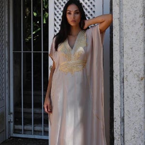 May include: A long, flowing, light beige kaftan with gold embroidery. The kaftan has a V-neck and short sleeves.
