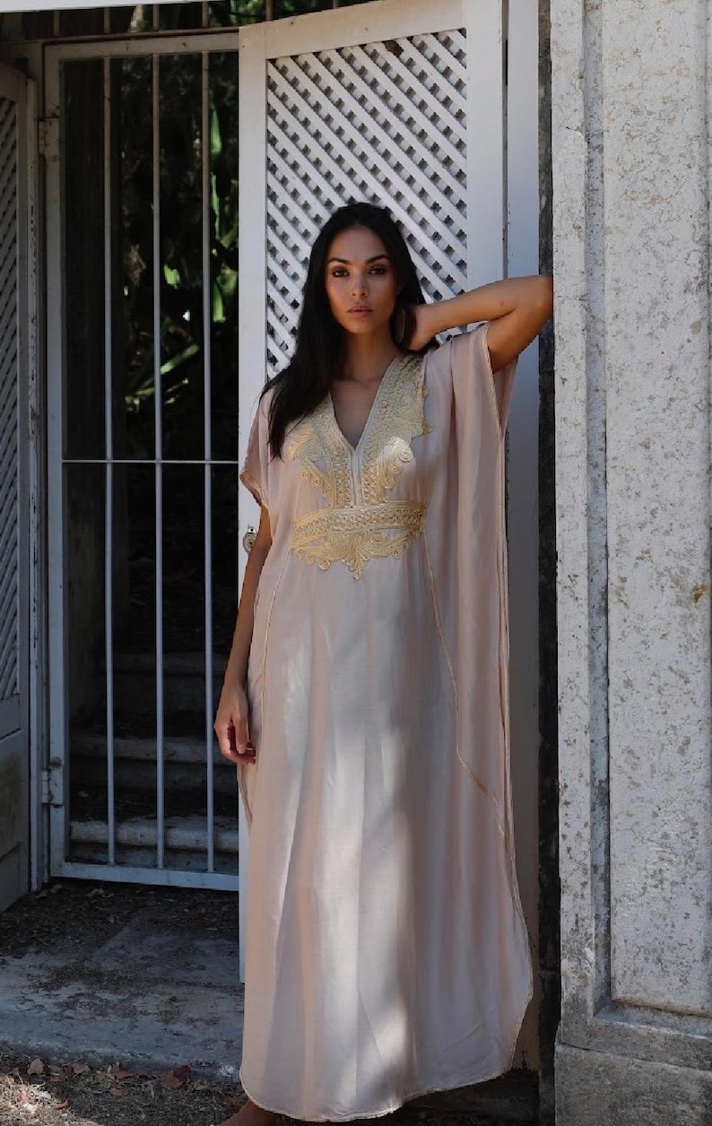 May include: A long, flowing, light beige kaftan with gold embroidery. The kaftan has a V-neck and short sleeves.
