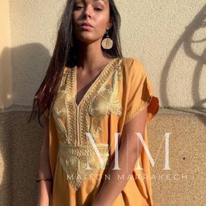 May include: A woman wearing a long, mustard yellow kaftan with gold embroidery. The kaftan has a V-neckline and short sleeves. The woman is wearing a gold bracelet on her left wrist and a large, round, gold earring on her left ear. The text "MM Maison Marrakech" is visible at the bottom of the image.