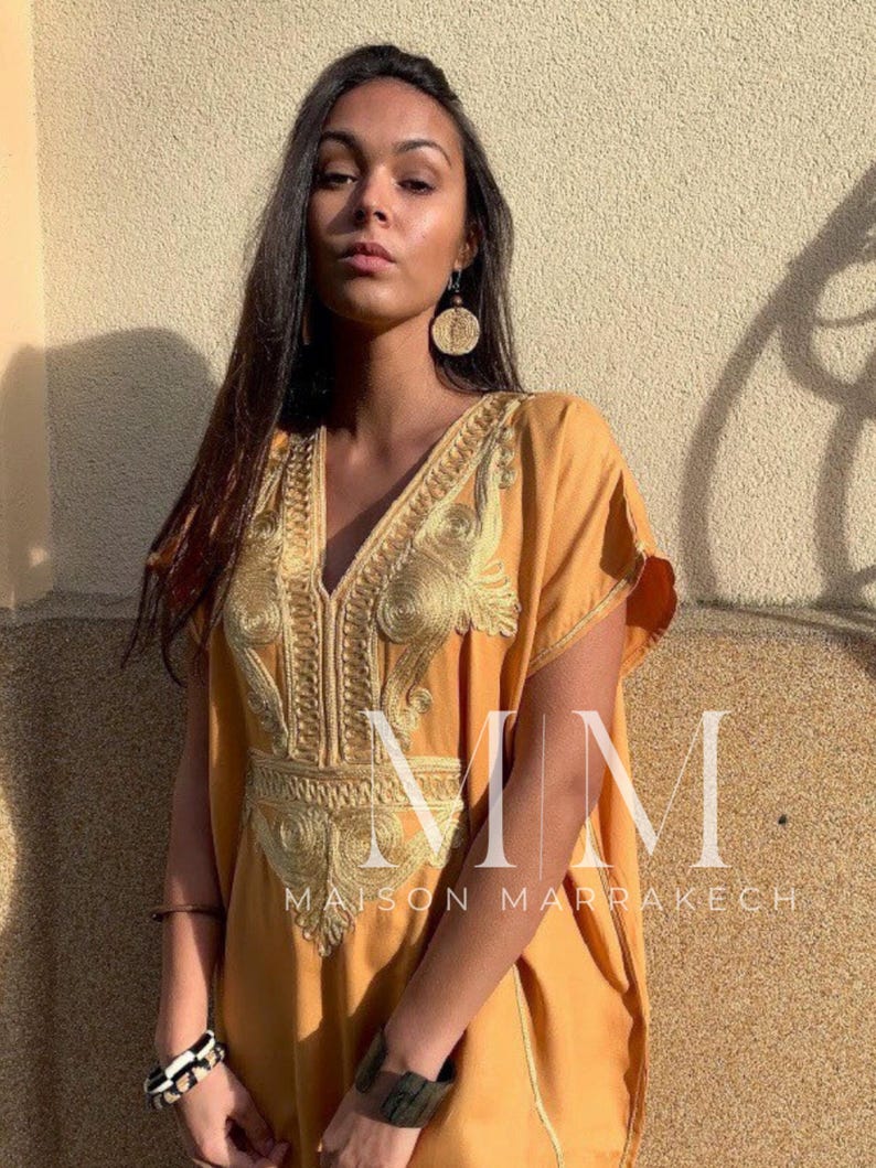 May include: A woman wearing a long, mustard yellow kaftan with gold embroidery. The kaftan has a V-neckline and short sleeves. The woman is wearing a gold bracelet on her left wrist and a large, round, gold earring on her left ear. The text "MM Maison Marrakech" is visible at the bottom of the image.