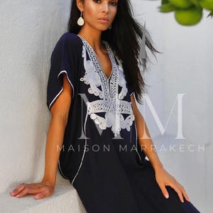 May include: A woman wearing a navy blue kaftan with white embroidery. The kaftan has a V-neck and short sleeves. The embroidery is intricate and features a floral design. The woman is standing in front of a white wall. The text "Maison Marrakech" is visible on the kaftan.