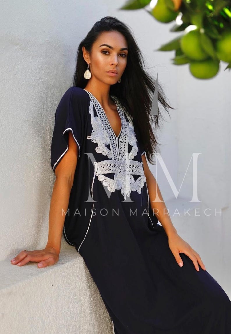 May include: A woman wearing a navy blue kaftan with white embroidery. The kaftan has a V-neck and short sleeves. The embroidery is intricate and features a floral design. The woman is standing in front of a white wall. The text "Maison Marrakech" is visible on the kaftan.