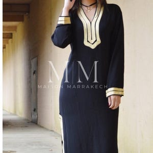 Puede incluir: Un vestido negro de manga larga con ribetes dorados alrededor del escote, las mangas y el dobladillo. El vestido tiene un escote en V y una abertura lateral. El texto "MM Maison Marrakech" est&aacute; impreso en el vestido.