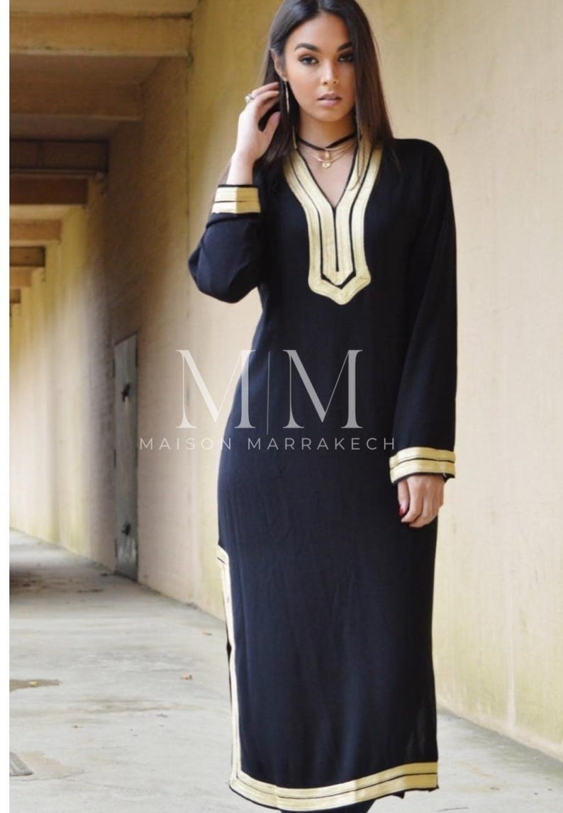 Puede incluir: Un vestido negro de manga larga con ribetes dorados alrededor del escote, las mangas y el dobladillo. El vestido tiene un escote en V y una abertura lateral. El texto "MM Maison Marrakech" est&aacute; impreso en el vestido.