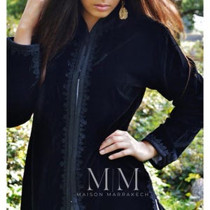 May include: A black velvet jacket with intricate embroidery along the front and sleeves. The jacket has a high collar and a long, flowing silhouette. The text "MM MAISON MARRAKECH" is visible at the bottom of the image.