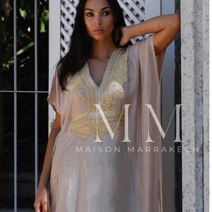 May include: A woman wearing a long, flowing beige kaftan with gold embroidery. The kaftan has a V-neckline and short sleeves. The woman is standing in front of a white wall with a lattice pattern. The text "MM Maison Marrakech" is visible on the kaftan.