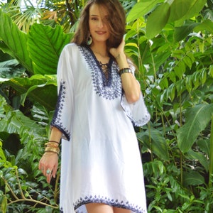 May include: White embroidered tunic dress with a lace-up neckline and blue embroidery. The dress is loose-fitting and has a flowy silhouette.