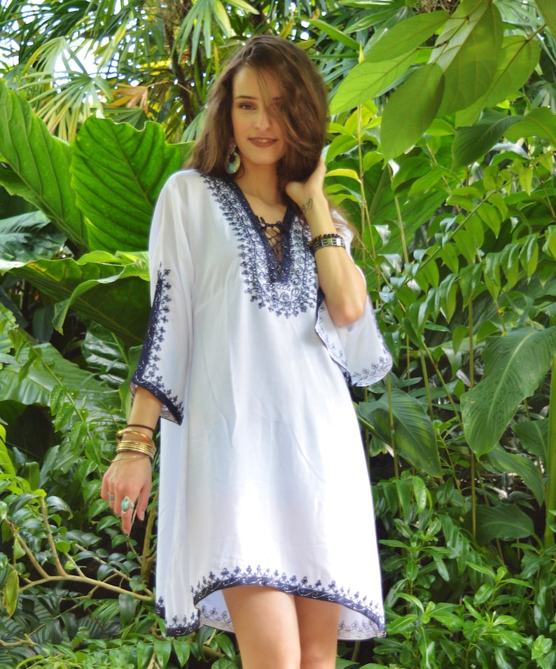 May include: White embroidered tunic dress with a lace-up neckline and blue embroidery. The dress is loose-fitting and has a flowy silhouette.
