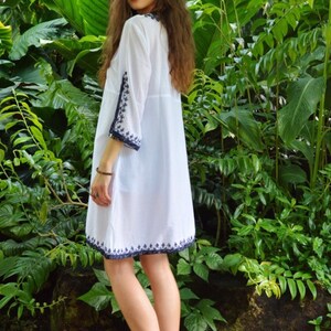 May include: A white cotton dress with blue embroidery around the neckline, sleeves, and hem. The dress is loose-fitting and has a V-neckline. The model is wearing the dress in a garden setting.
