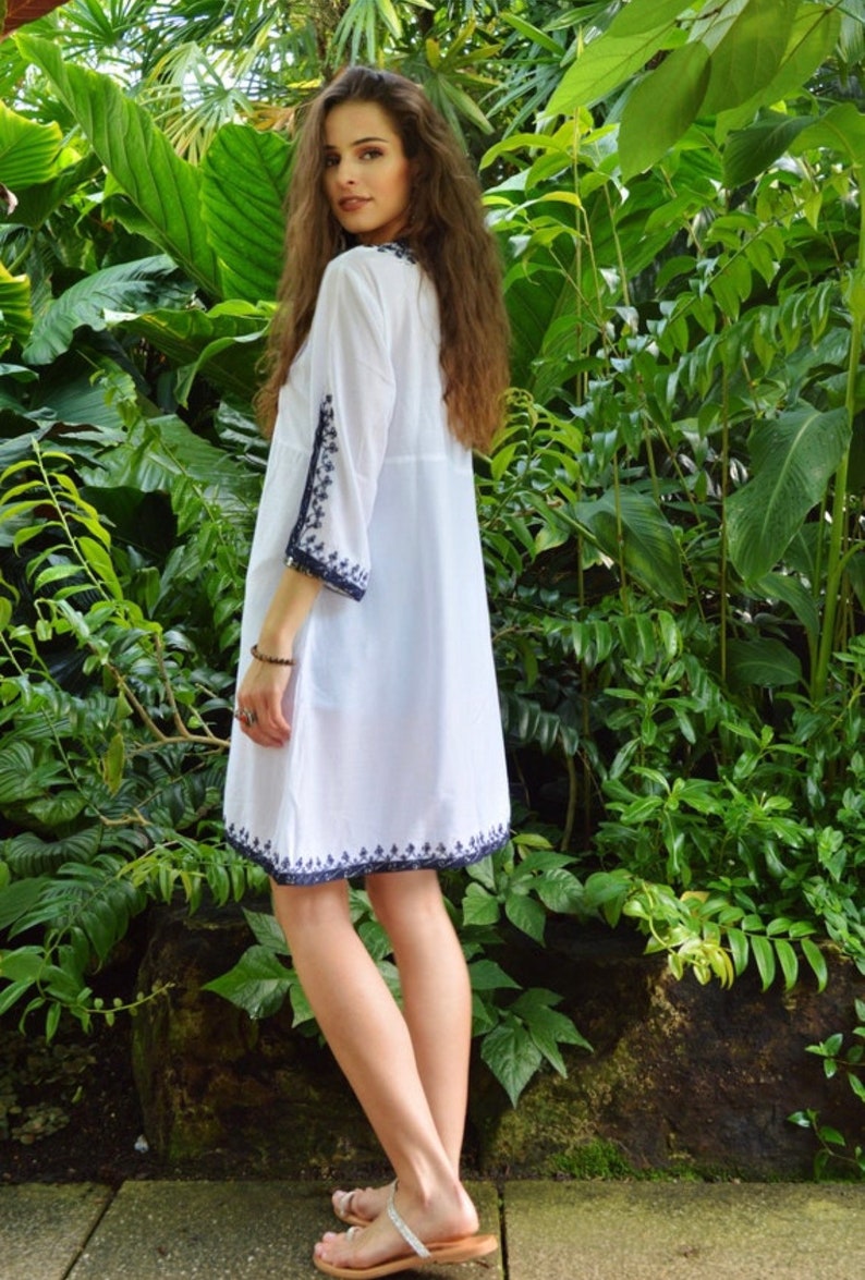 May include: A white cotton dress with blue embroidery around the neckline, sleeves, and hem. The dress is loose-fitting and has a V-neckline. The model is wearing the dress in a garden setting.