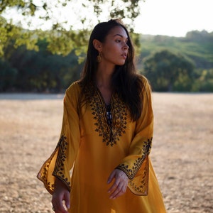 May include: A woman wearing a yellow tunic with black embroidery. The tunic has long sleeves and a V-neckline. The woman is standing in a field with trees in the background.