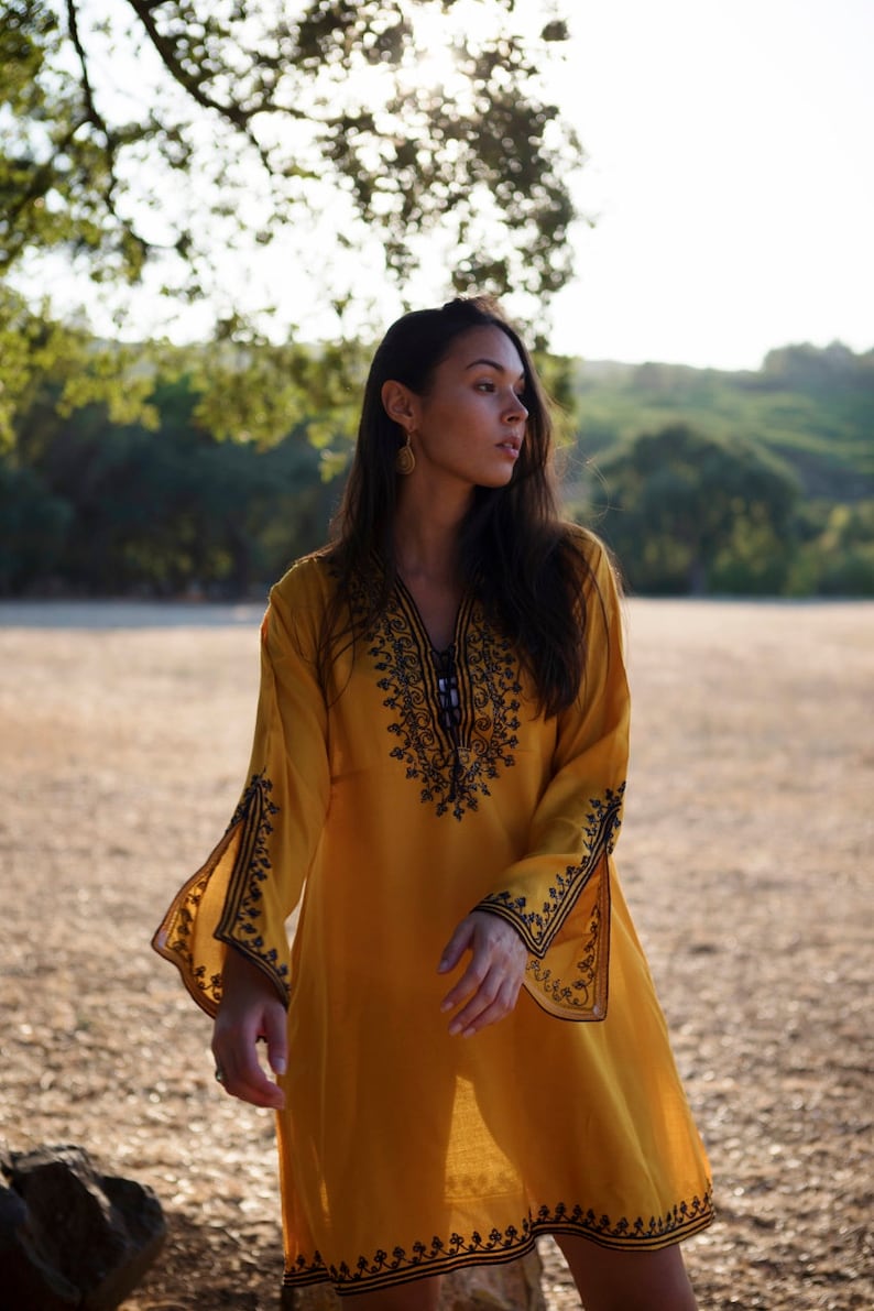 May include: A woman wearing a yellow tunic with black embroidery. The tunic has long sleeves and a V-neckline. The woman is standing in a field with trees in the background.