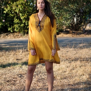 May include: A woman wearing a yellow embroidered dress with long sleeves and a V-neck. She is standing in a field of dry grass.