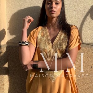 May include: A woman wearing a long, loose-fitting, mustard yellow kaftan with gold embroidery. The kaftan has a V-neckline and short sleeves. The woman is standing in front of a wall with the text "MMM Maison Marrakech" printed on it.