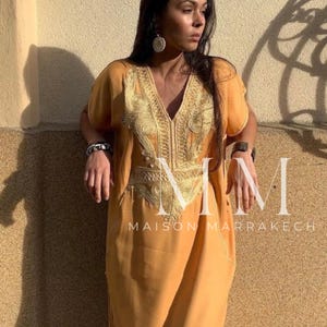 May include: A long, loose-fitting, mustard yellow kaftan with gold embroidery. The kaftan has a V-neck and short sleeves. The embroidery is intricate and features a floral design. The text "Maison Marrakech" is visible at the bottom of the image.