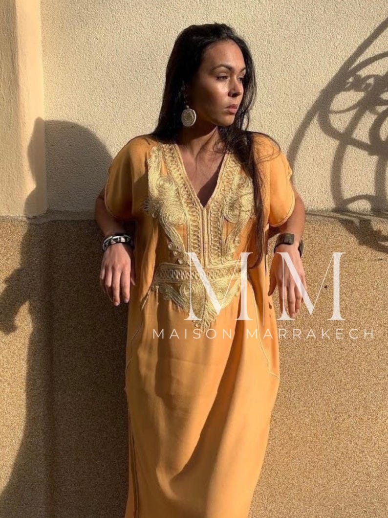 May include: A long, loose-fitting, mustard yellow kaftan with gold embroidery. The kaftan has a V-neck and short sleeves. The embroidery is intricate and features a floral design. The text "Maison Marrakech" is visible at the bottom of the image.