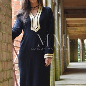 Puede incluir: Un vestido negro de manga larga con ribetes dorados alrededor del cuello, las mangas y el dobladillo. El vestido tiene cuello en V y est&aacute; bordado con un patr&oacute;n geom&eacute;trico. El texto "MMM Maison Marrakech" es visible en el vestido.