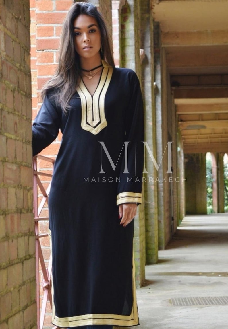 Puede incluir: Un vestido negro de manga larga con ribetes dorados alrededor del cuello, las mangas y el dobladillo. El vestido tiene cuello en V y est&aacute; bordado con un patr&oacute;n geom&eacute;trico. El texto "MMM Maison Marrakech" es visible en el vestido.