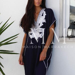May include: A navy blue kaftan with white embroidery. The kaftan has a V-neckline and short sleeves. The embroidery is intricate and features a floral design. The kaftan is long and flowing. The text "Maison Marrakech" is visible on the bottom of the image.