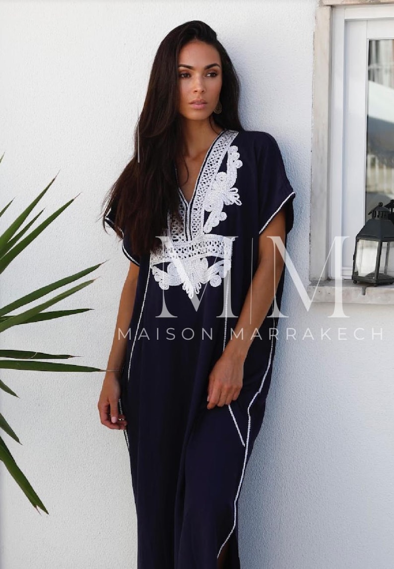 May include: A navy blue kaftan with white embroidery. The kaftan has a V-neckline and short sleeves. The embroidery is intricate and features a floral design. The kaftan is long and flowing. The text "Maison Marrakech" is visible on the bottom of the image.