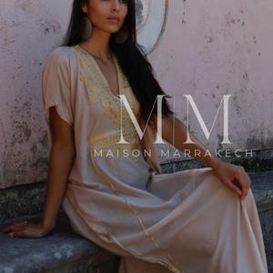 May include: A woman wearing a long, light beige kaftan with gold embroidery and trim. The kaftan has a V-neckline and short sleeves. The woman is sitting on a stone ledge with a pink wall behind her. The text "MM MAISON MARRAKECH" is visible in the image.