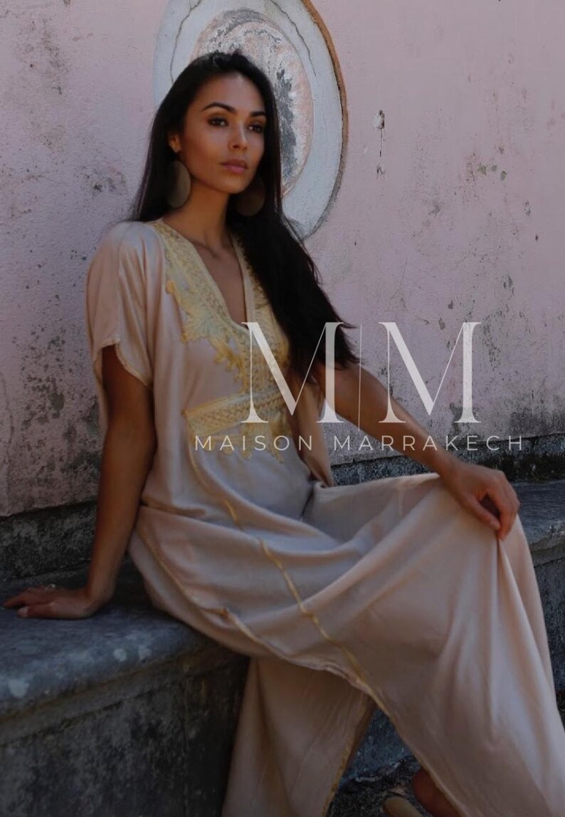 May include: A woman wearing a long, light beige kaftan with gold embroidery and trim. The kaftan has a V-neckline and short sleeves. The woman is sitting on a stone ledge with a pink wall behind her. The text "MM MAISON MARRAKECH" is visible in the image.