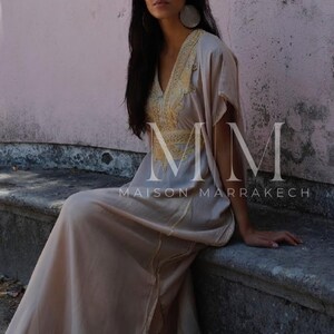 May include: A woman wearing a long, flowing beige dress with gold embroidery. The dress has a V-neckline and short sleeves. The woman is sitting on a stone bench. The text "MM Maison Marrakech" is visible on the dress.