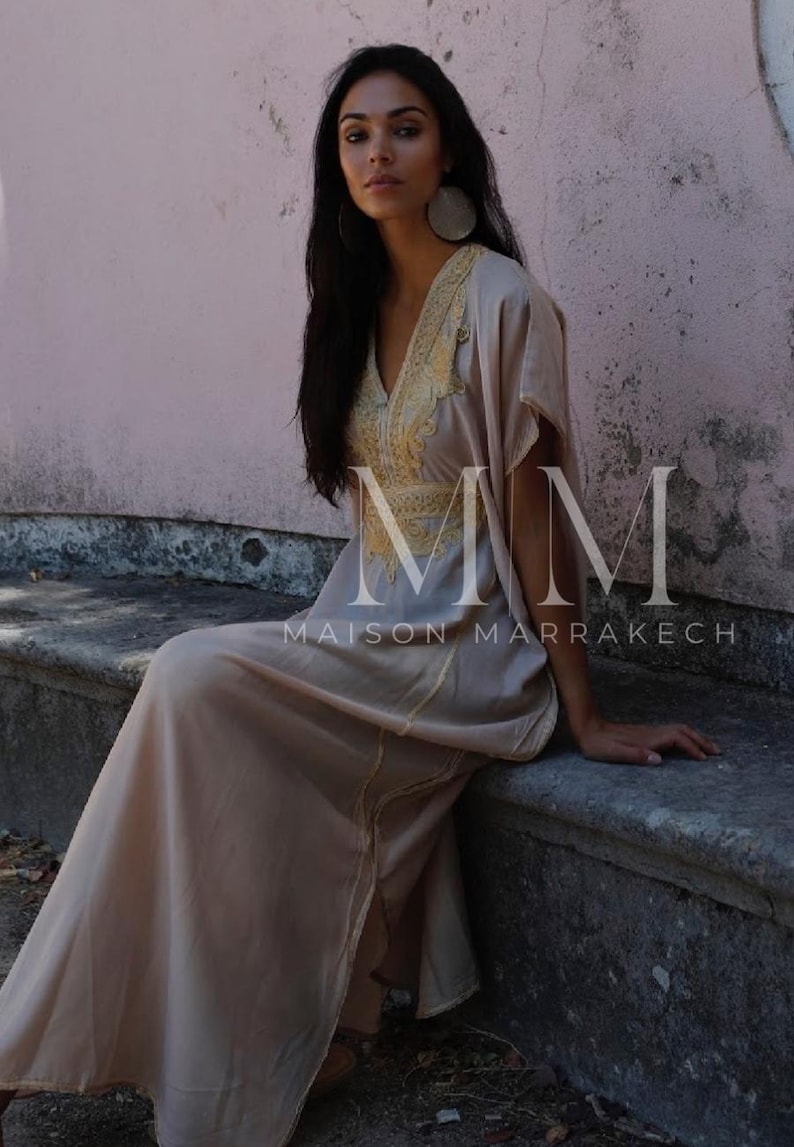 May include: A woman wearing a long, flowing beige dress with gold embroidery. The dress has a V-neckline and short sleeves. The woman is sitting on a stone bench. The text "MM Maison Marrakech" is visible on the dress.