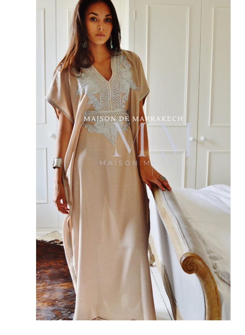Puede incluir: Un caft&aacute;n largo beige, con cuello en V y mangas cortas. El vestido presenta un intrincado bordado blanco alrededor del escote y el corpi&ntilde;o. El texto "MAISON DE MARRAKECH" es visible en el vestido.