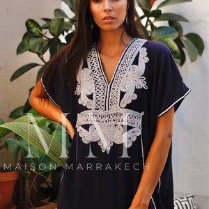 May include: A navy blue kaftan with white embroidery. The embroidery features a floral design and is accented with silver thread. The kaftan has a V-neckline and short sleeves. The text "MAISON MARRAKECH" is visible at the bottom of the image.